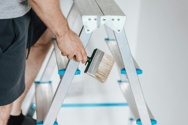 A man wearing black shorts stands on a ladder with a paintbrush in his hand.